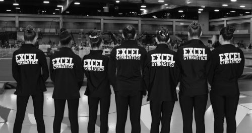 Four gymnasts wearing 'Excel Gymnastics' jackets stand with their backs to the camera.
