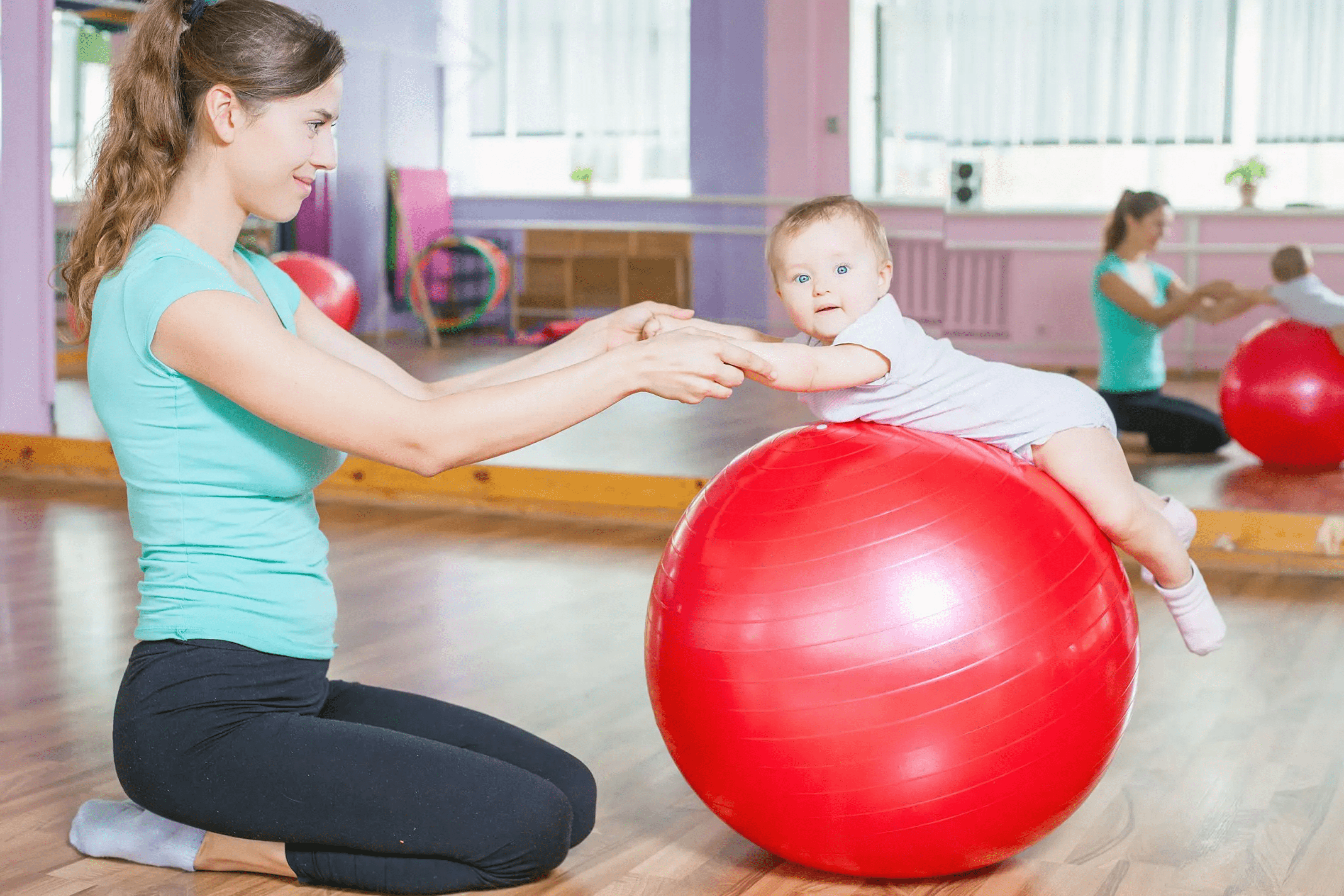 A woman helping a baby balance on a large red exercise ball.