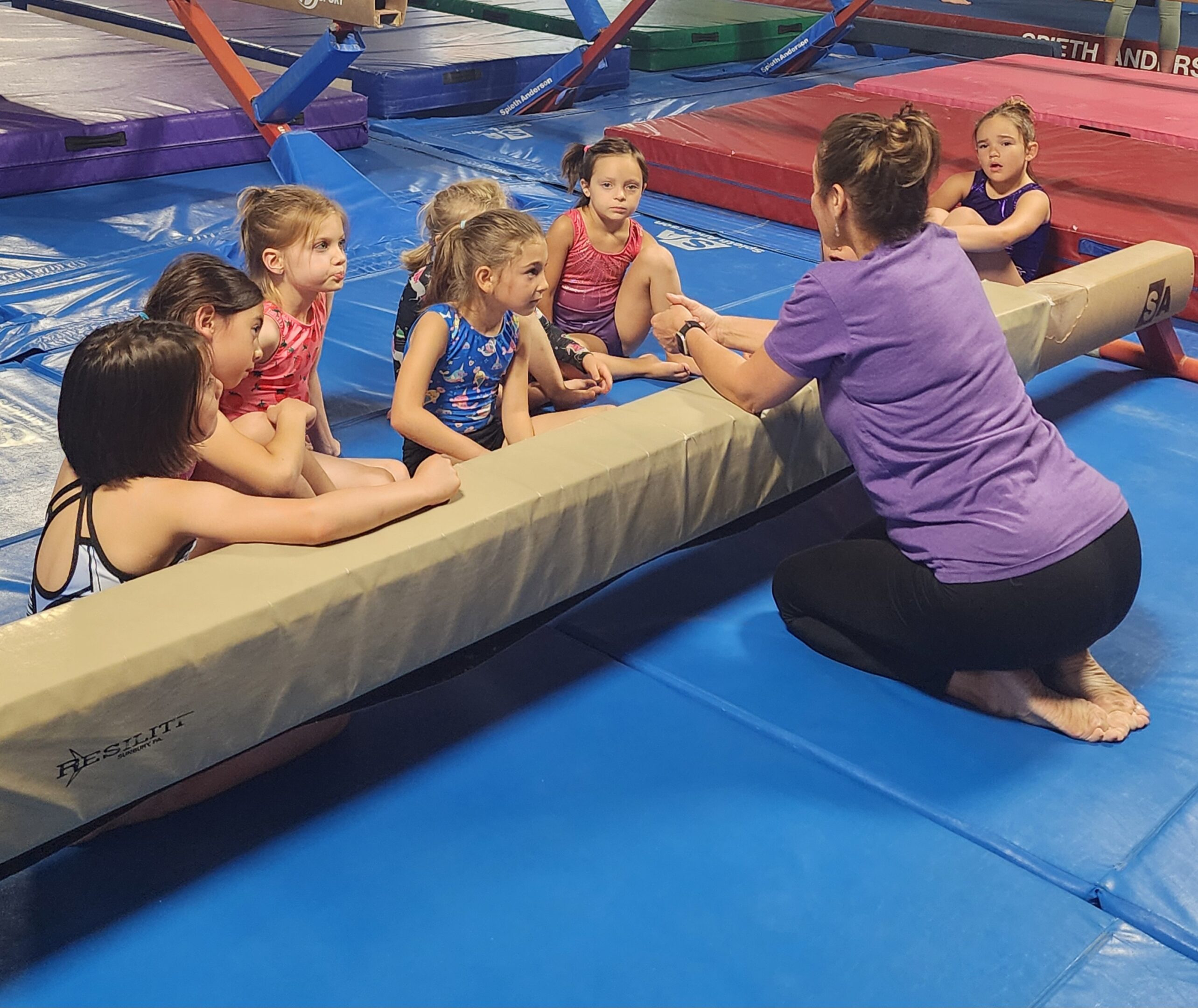 Children sitting on a gym mat listening to an instructor.