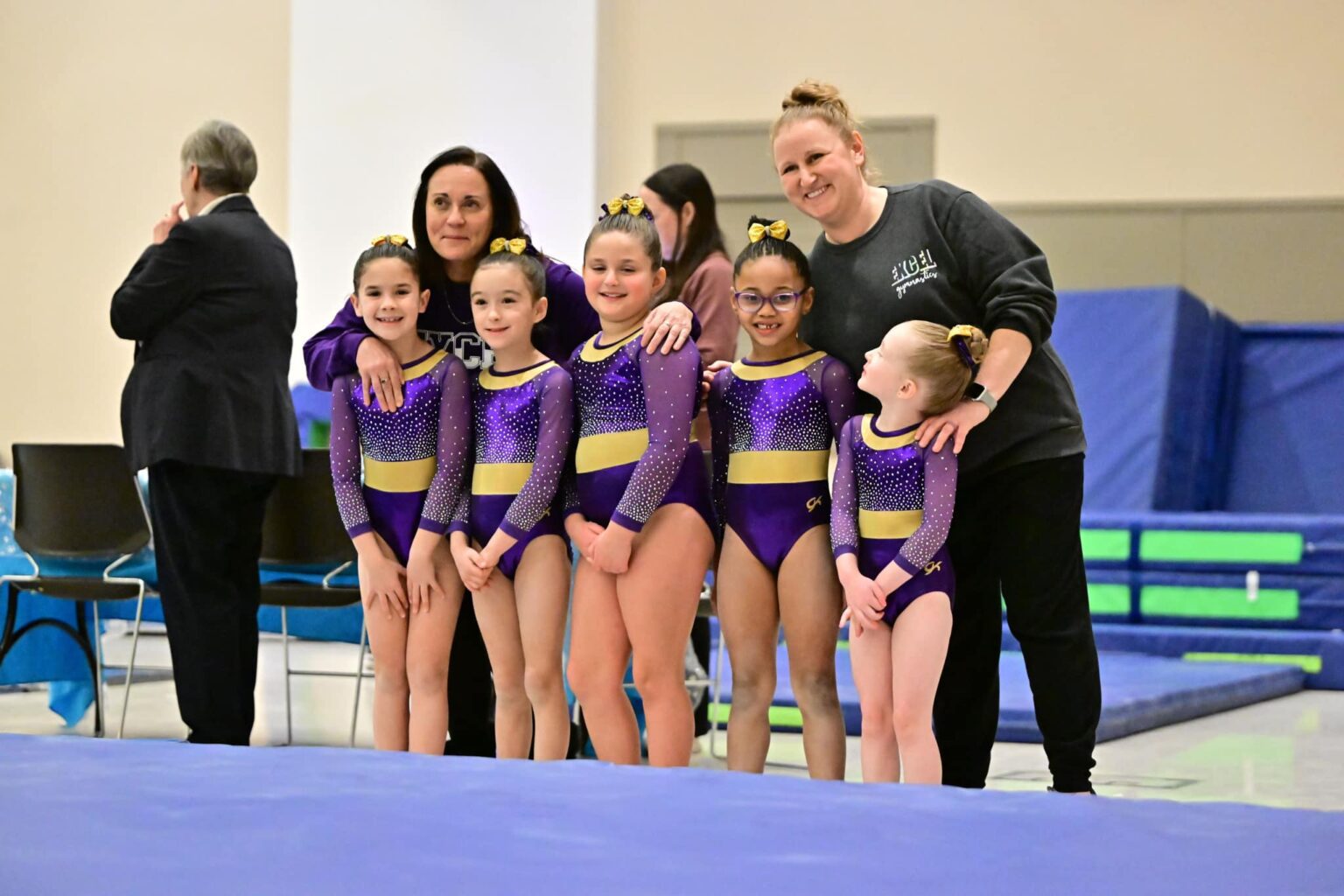 A group of young gymnasts in purple and yellow leotards with their coaches.